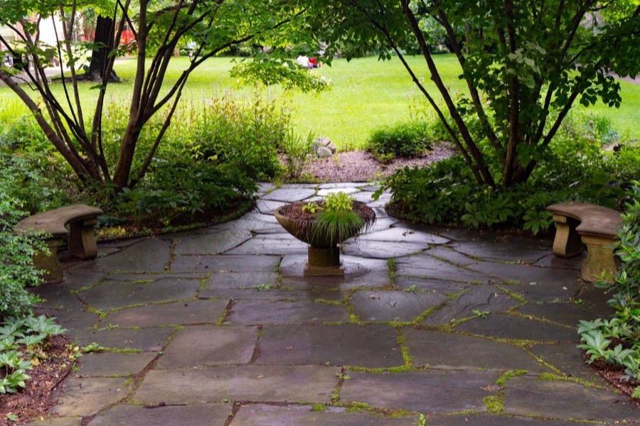 A flagstone terrace shaded by multi-stem trees, stone benches at its edges and a planted urn at its center, opening onto a mown lawn