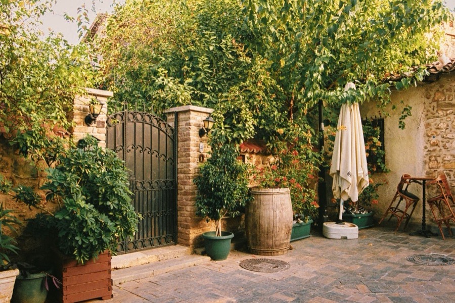 A walled courtyard paved wall to wall in stone: an iron gate between brick piers, potted trees and timber planters, lanterns in warm evening light